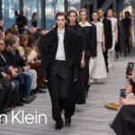 Models walking the runway at a Calvin Klein fashion show, showcasing minimalist black and neutral-toned outfits in front of a seated audience.