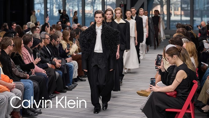Models walking the runway at a Calvin Klein fashion show, showcasing minimalist black and neutral-toned outfits in front of a seated audience.